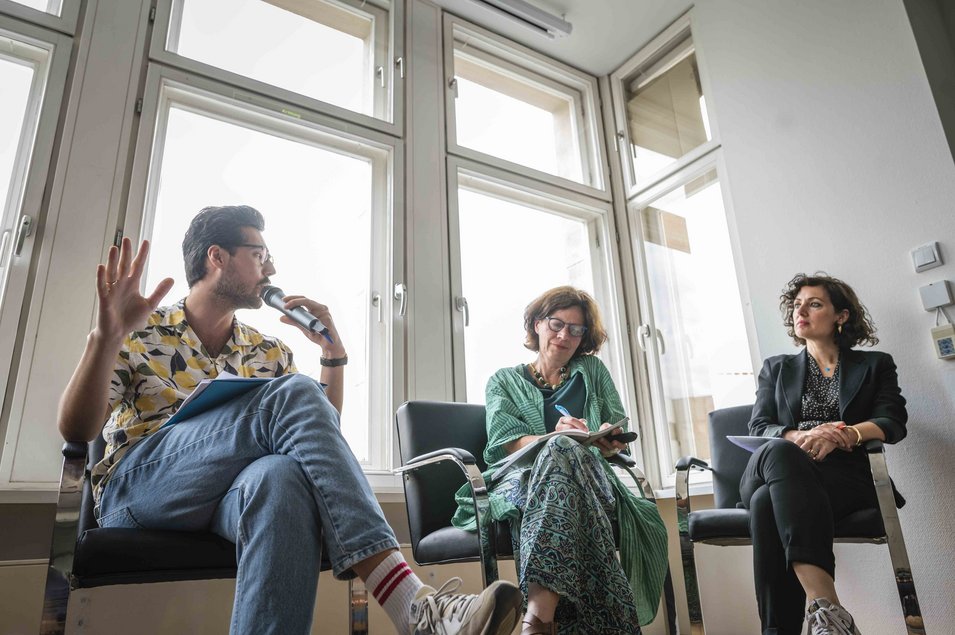 Ozan Zakariya Keskinkılıç spricht bei der DeZIM-Lunch Discussion in ein Mikrofon. Neben ihm sitzen Karen Phalet und Naika Foroutan, die ihm zuhören. Das Foto hat Paul Lovis Wagner gemacht.