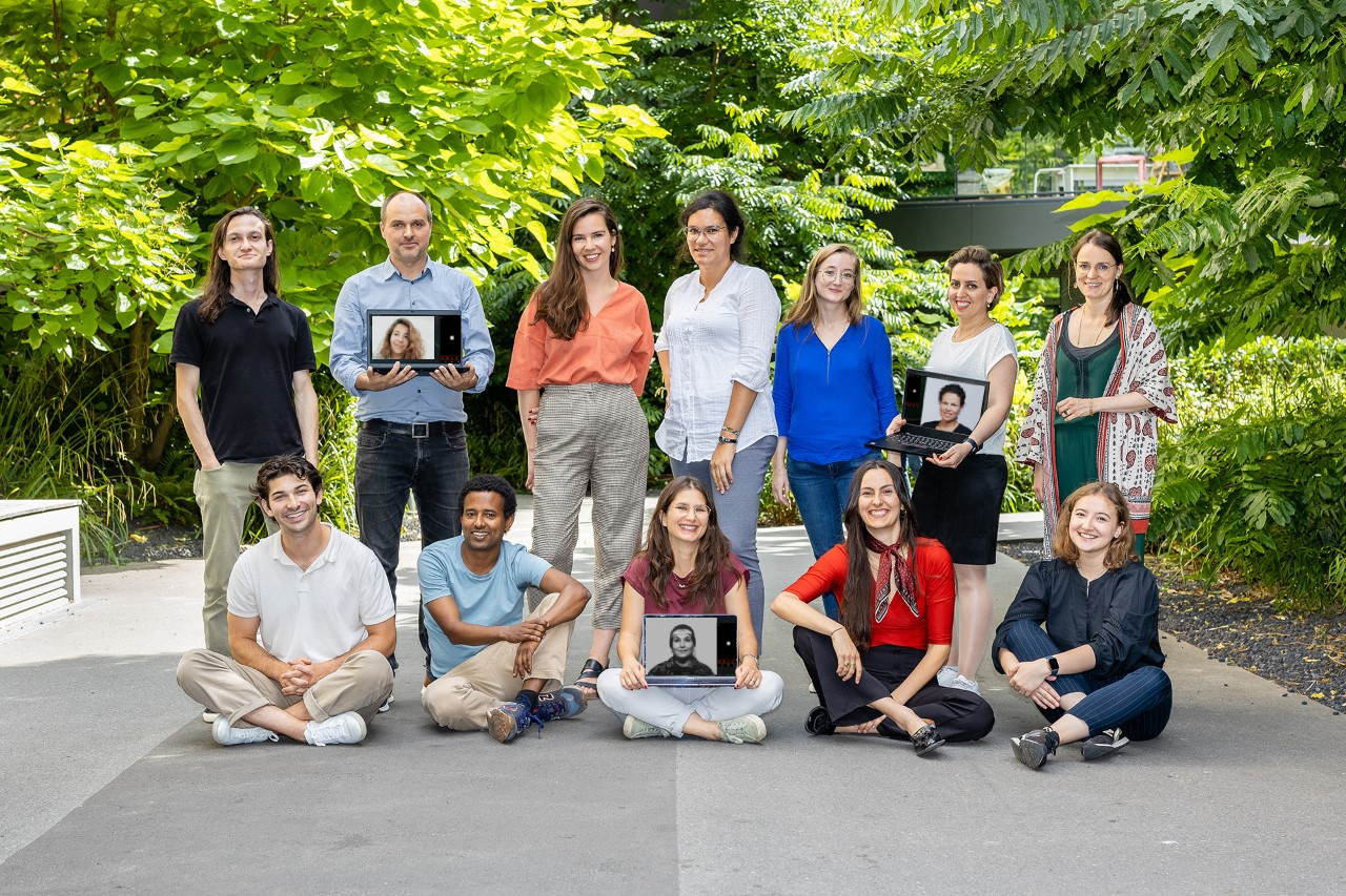 The photo shows a total of twelve employees of the Migration Department of the DeZIM Institute. Seven people are standing behind five people sitting on a gray floor. All are wearing summer clothes. Trees and bushes can be seen in the background. The photo was taken by Ines Grabner in July 2022.